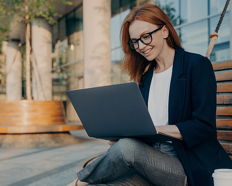 Eine lächelnde Frau mit roten Haaren sitzt auf einer Holzbank und arbeitet an einem Laptop. Sie trägt eine schwarze Jacke und eine weiße Bluse. Neben ihr steht ein Kaffeebecher. Der Hintergrund zeigt eine moderne Architektur.
