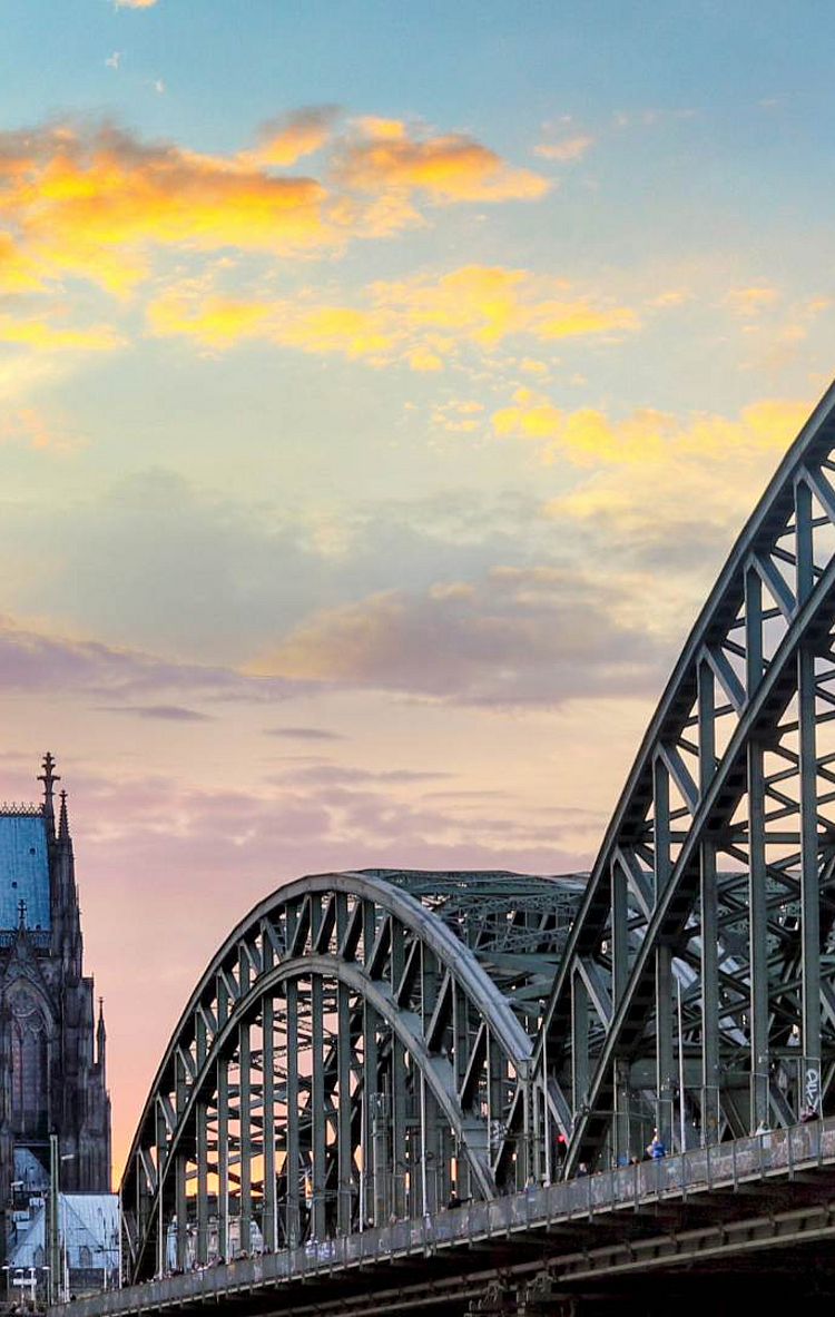 Ein Blick auf den Kölner Dom mit seinen markanten Türmen vor einem bunten Sonnenuntergang. Im Vordergrund ist eine Brücke sichtbar, die über einen Fluss führt, und verbindet die beiden Ufer der Stadt.